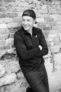Photo of guy with ball cap smiling while leaning against brick wall.